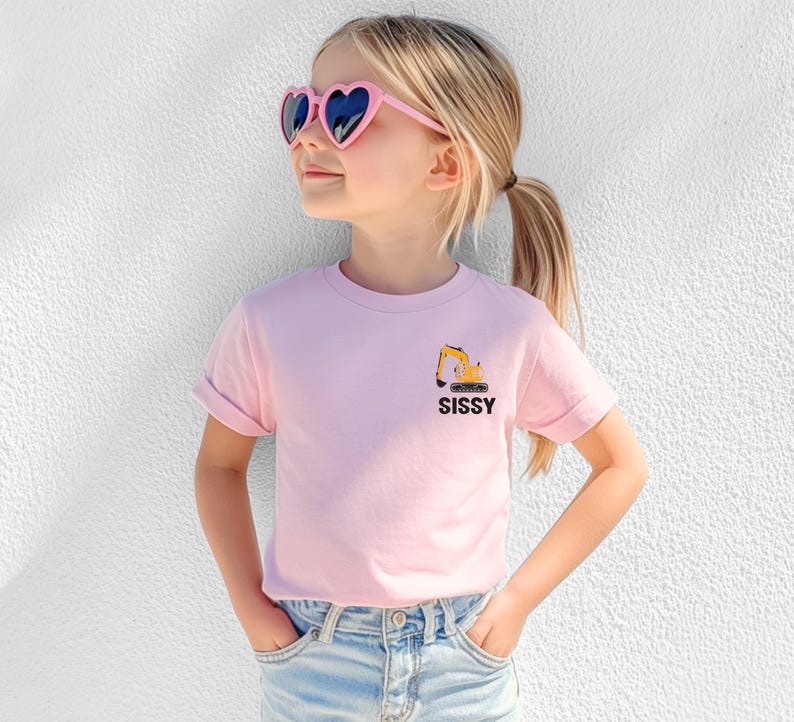 May include: A light pink short-sleeved t-shirt featuring a small graphic of a yellow excavator on the left chest area above the black text SISSY. The shirt is paired with light blue denim jeans and pink heart-shaped sunglasses.