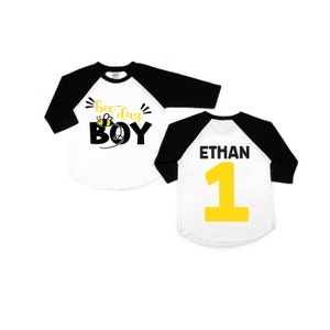 May include: A white and black baseball style shirt with a bee and the text "bee-day BOY" on the front. The back of the shirt has the name "ETHAN" and the number "1" in yellow.