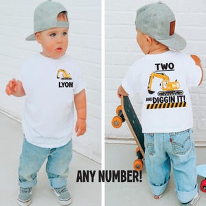 May include: A white t-shirt featuring a yellow excavator graphic and the name LYON on the front, with a larger yellow excavator graphic, the text TWO AND DIGGIN IT!, and a black and yellow caution stripe pattern printed on the back of the shirt.