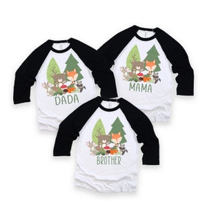 May include: Three white baseball shirts with black sleeves. The shirts feature a cartoon image of a bear and fox in a forest setting. The shirts are labeled "Dada", "Mama", and "Brother".