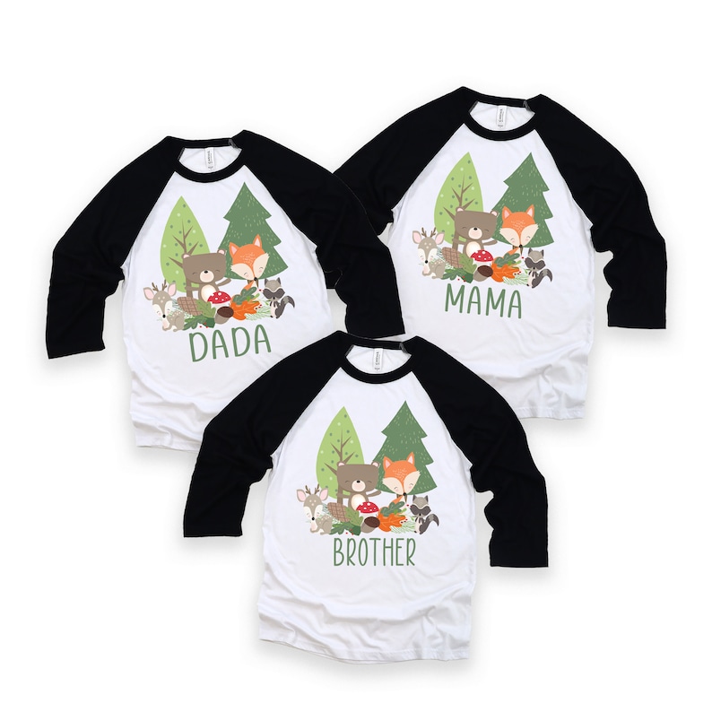 May include: Three white baseball shirts with black sleeves. The shirts feature a cartoon image of a bear and fox in a forest setting. The shirts are labeled "Dada", "Mama", and "Brother".