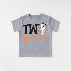Pu&ograve; includere: Una maglietta grigia a maniche corte con le parole "TWo Spooky" in nero e arancione. Un fantasma bianco con un cappello nero e dettagli arancioni &egrave; a destra del testo. Piccoli pipistrelli neri e dettagli arancioni completano il design di Halloween.