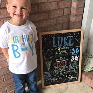 May include: A chalkboard sign with the name "Luke" written in blue and pink. The sign lists his age, weight, height, and favourite things. The sign is decorated with colourful sprinkles and a drawing of an ice cream cone. A young boy wearing a white t-shirt with the words "Birthday Boy" and a blue and yellow ice cream cone graphic is standing next to the sign.