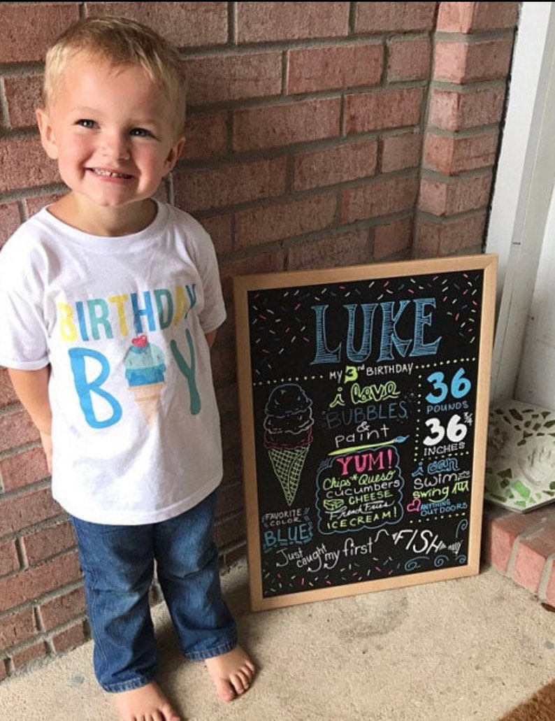 May include: A chalkboard sign with the name "Luke" written in blue and pink. The sign lists his age, weight, height, and favourite things. The sign is decorated with colourful sprinkles and a drawing of an ice cream cone. A young boy wearing a white t-shirt with the words "Birthday Boy" and a blue and yellow ice cream cone graphic is standing next to the sign.