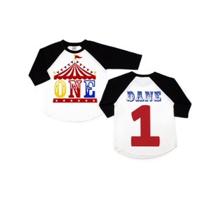 Puede incluir: Dos camisetas de b&eacute;isbol blancas y negras con un gr&aacute;fico de carpa de circo roja y amarilla en la parte delantera. Una camiseta dice "ONE" y la otra dice "DANE 1".
