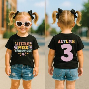 May include: A black short-sleeve t-shirt featuring the text 'LITTLE MISS THREENAGER' with colorful flower graphics on the front and the name 'AUTUMN' above a large number '3' in light pink on the back, paired with blue denim shorts.