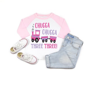 May include: A white and pink toddler's t-shirt with a pink train and the text "Chugga Chugga Three Three!" printed on the front. The shirt has a pink and white baseball style design.