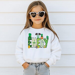 May include: White sweatshirt with a green, gold, and white design of leprechauns and shamrocks spelling out the name "EMERY". A young girl is wearing the sweatshirt and has on heart-shaped sunglasses.