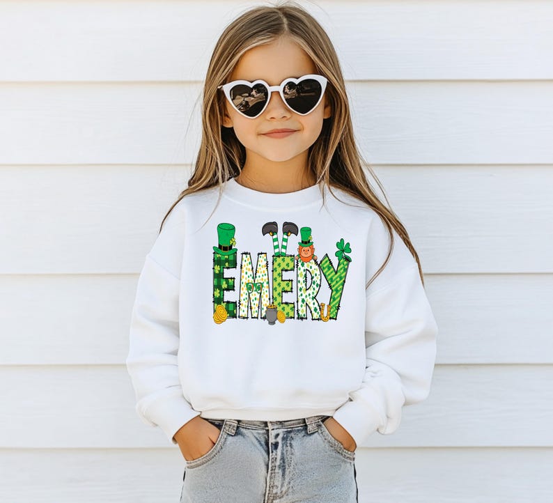 May include: White sweatshirt with a green, gold, and white design of leprechauns and shamrocks spelling out the name "EMERY". A young girl is wearing the sweatshirt and has on heart-shaped sunglasses.