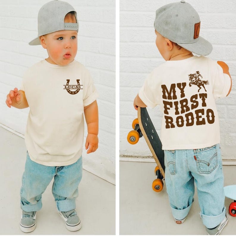 Op de afbeelding: Een peuter draagt een cr&egrave;mekleurig T-shirt met de tekst "MY FIRST RODEO" in bruin op de rug en een hoefijzerontwerp met "LUCAS" op de voorkant. Het kind draagt een lichtblauwe spijkerbroek en een grijze baseballcap.
