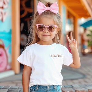 May include: A white short-sleeved t-shirt featuring the word SISTER printed in a colorful floral pattern on the left chest, paired with a large light pink hair bow and light pink sunglasses with dark lenses, worn with blue denim jeans.