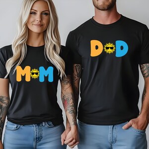 May include: Two black short-sleeved t-shirts. One shirt features the text MOM in orange and blue with a yellow sun wearing sunglasses as the letter O. The other shirt features the text DAD in orange and blue with a yellow sun wearing sunglasses as the letter A.