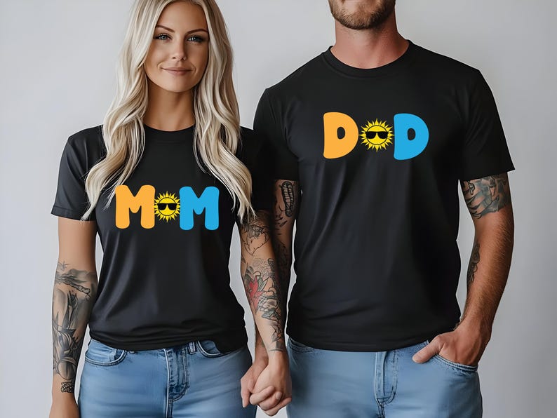 May include: Two black short-sleeved t-shirts. One shirt features the text MOM in orange and blue with a yellow sun wearing sunglasses as the letter O. The other shirt features the text DAD in orange and blue with a yellow sun wearing sunglasses as the letter A.