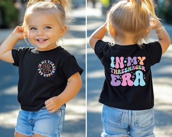 May include: Black short-sleeved t-shirt with a rainbow graphic that says "In My Three-nager Era" on the back and a rainbow graphic with the number three and the text "In My Three-nager Era" on the front.