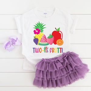 May include: White t-shirt with a colorful fruit design and the text "TWO-tti FRUTTI". The shirt is paired with a purple tulle skirt.