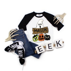 May include: A Halloween-themed outfit featuring a white and black raglan shirt with a ghost and pumpkin graphic, the name "MASON", and the word "EEK". The outfit includes blue jeans and black sneakers. Decorative skeleton hands are also present.