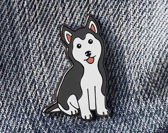 May include: A black and white enamel pin featuring a cartoon husky dog with a happy expression. The pin is sitting on a blue denim background.