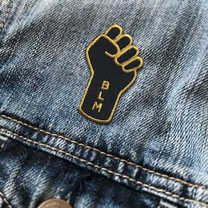 May include: Black fist raised in the air with the letters BLM in gold embroidery on a denim jacket.