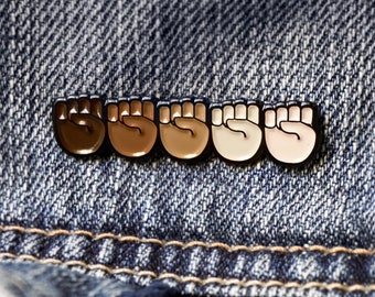 May include: A horizontal enamel pin featuring five raised fists arranged in a row, each rendered in a different skin tone ranging from dark brown to light beige, all outlined in black metal, attached to a piece of blue denim fabric.