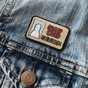May include: A beige and brown embroidered patch with the text "SIMP CARD" in red and white lettering. The patch features a white silhouette of a person's head in a light blue rectangle.