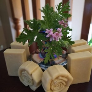 Puede incluir: Una colección de jabones hechos a mano con diseño de rosa. Los jabones son de color beige claro y están dispuestos en círculo alrededor de un pequeño jarrón con una planta verde y flores rosas.