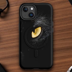 Peut inclure: Coque de téléphone noire avec un gros plan sur un œil de chat au iris jaune. Le boîtier a un design épuré et un anneau magnétique à l'arrière. La coque est posée sur une surface en bois, entourée d'une montre, de lunettes et d'un carnet.