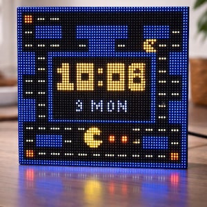 Op de afbeelding: Een blauwe digitale klok in pixel art die de tijd 10:06, de dag van de week en een Pac-Man-achtige afbeelding weergeeft. De klok heeft een zwarte rand en staat op een houten oppervlak.