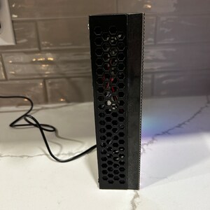 May include: A black rectangular electronic device with a honeycomb pattern on one side and a series of small holes on the other. A black cable extends from the device. The device is on a white surface.
