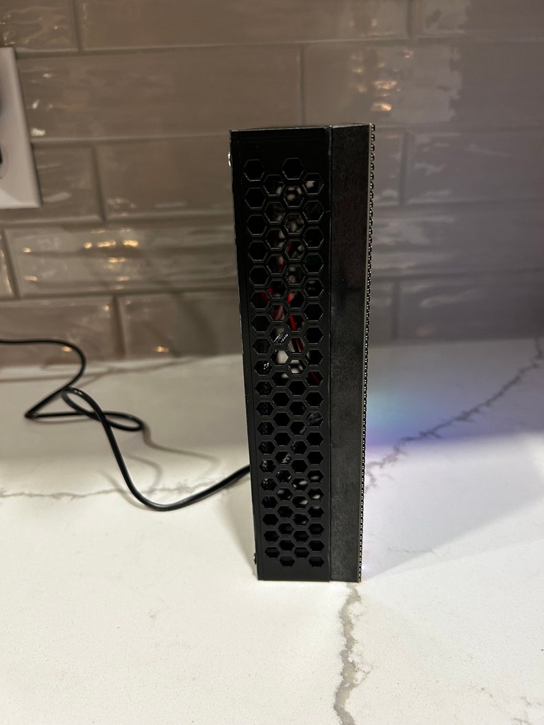 May include: A black rectangular electronic device with a honeycomb pattern on one side and a series of small holes on the other. A black cable extends from the device. The device is on a white surface.