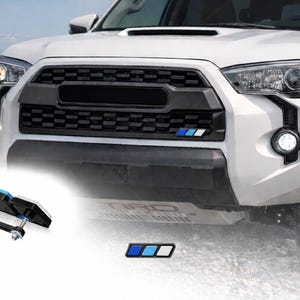 May include: White SUV with a black grille and blue, white, and black emblem. A black and blue accessory is in the foreground. The vehicle has fog lights and a mountain range in the background.