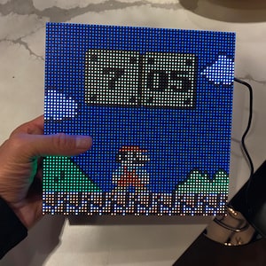 May include: A square LED display showing a pixelated image of Super Mario Bros. The display features Mario, the text "MGS", and a blue sky with green hills. The display is held by a hand. The display is connected to a black cable.