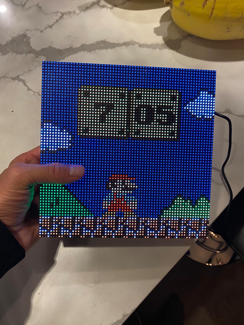 May include: A square LED display showing a pixelated image of Super Mario Bros. The display features Mario, the text "MGS", and a blue sky with green hills. The display is held by a hand. The display is connected to a black cable.