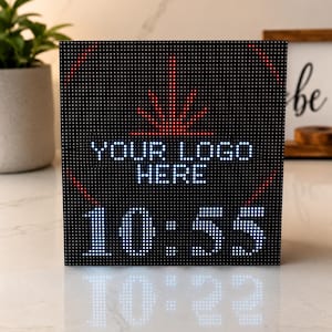以下が含まれることがあります： 「YOUR LOGO HERE」の文字と10:55の時刻が白く表示された黒いLEDディスプレイキューブ。上部には赤いグラフィックがあります。キューブは白い表面に置かれています。