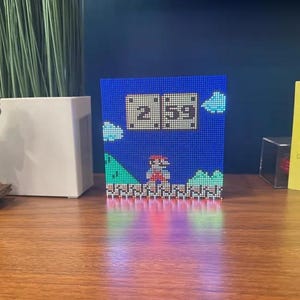 May include: A blue pixel art clock showing the time 2:59, with a Super Mario Bros. design. The clock has a blue background with pixelated clouds, green hills, and a red-clad Mario character. The clock is on a wooden surface.