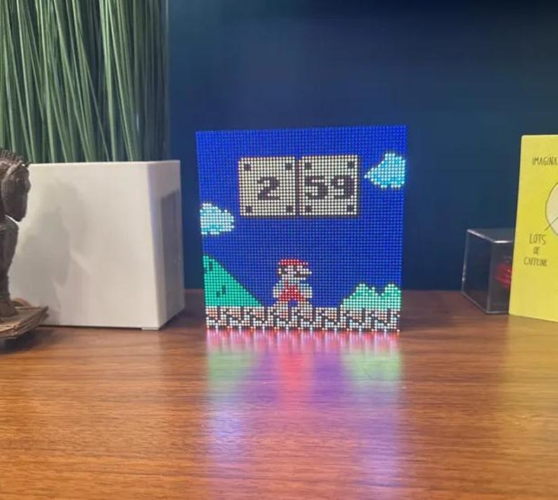 May include: A blue pixel art clock showing the time 2:59, with a Super Mario Bros. design. The clock has a blue background with pixelated clouds, green hills, and a red-clad Mario character. The clock is on a wooden surface.