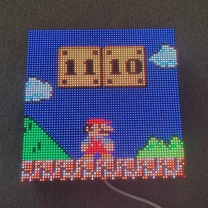 May include: Square LED display showing a pixelated scene from a classic video game. The scene includes a character in red overalls, green hills, and a blue sky with clouds. The display is lit with various colours.