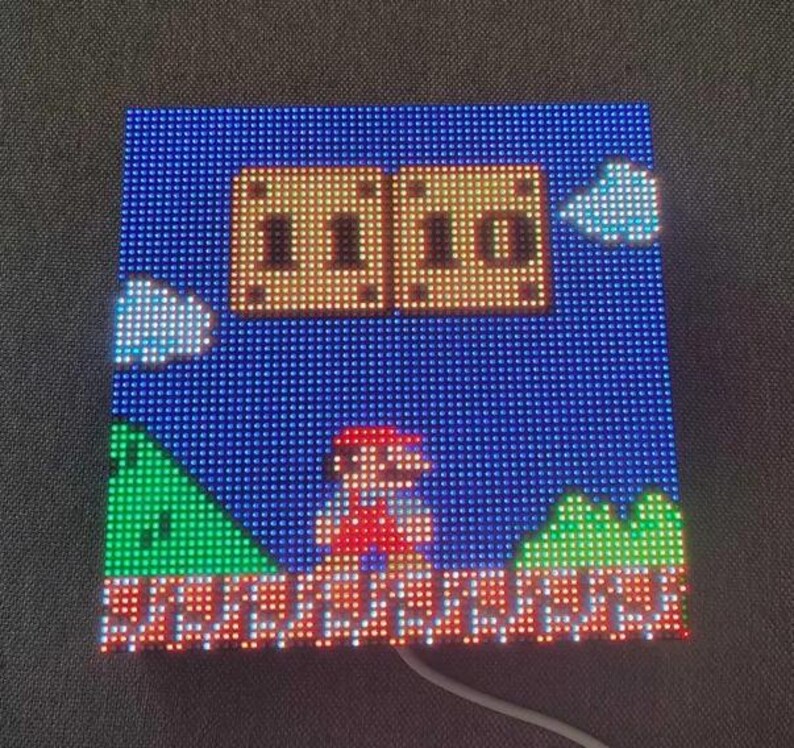 May include: Square LED display showing a pixelated scene from a classic video game. The scene includes a character in red overalls, green hills, and a blue sky with clouds. The display is lit with various colours.