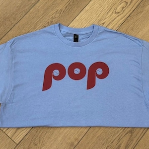 May include: A light blue t-shirt with the word "pop" in a bold, red font. The shirt is made of a soft material and is laid flat on a wooden surface. The design features a unique, stylized font.