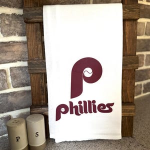 May include: White kitchen towel with a maroon "P" and "Phillies" logo. The towel is hanging on a wooden rack.