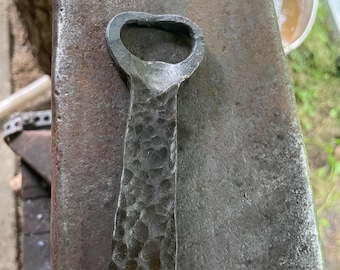 May include: A hand-forged bottle opener made of dark gray metal with a distinct hammered, textured finish on the handle and a rounded, open loop at the top for gripping bottle caps, showcasing a rugged, industrial appearance.
