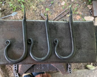 May include: Three dark gray metal hooks with a curved J-shape and a decorative curled end. Each hook features a flat, pointed top section with a small circular hole for mounting, showcasing a textured, hand-forged appearance on a flat surface.