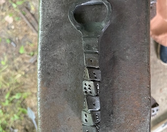 May include: A hand-forged dark gray metal bottle opener featuring a handle designed as a vertical stack of five individual dice. The metal has a textured, industrial finish with visible pips on each die face, leading to a rounded opening loop.