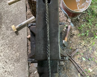 May include: A hand-forged iron fire poker featuring a long, twisted metal shaft and a circular loop handle at one end. The poker is resting vertically on top of a heavy, dark gray metal anvil, showcasing a textured, dark charcoal finish.