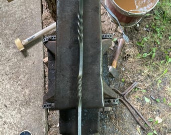 May include: A hand-forged iron fire poker featuring a long, slender shaft with two distinct twisted sections and a decorative scrolled loop handle at the end. The metal has a dark, matte finish and rests on a heavy, rectangular steel anvil.