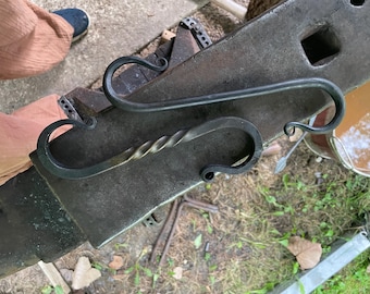 May include: A hand-forged black metal S-hook featuring a central twisted bar design with curled ends. The hook is crafted from dark iron and displays a textured, metallic finish consistent with traditional blacksmithing techniques and metalwork.