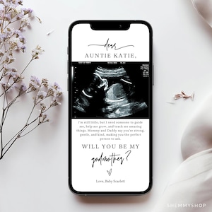 May include: A smartphone displays a baby's sonogram with text that reads "Dear Auntie Katie, Will you be my godmother?" The message is in black script font on a white background. The phone is surrounded by white flowers.