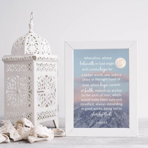 May include: A white framed print with a quote about faith and hope, set against a backdrop of a mountain range and a full moon. A decorative white lantern and lace ribbon are in the foreground.