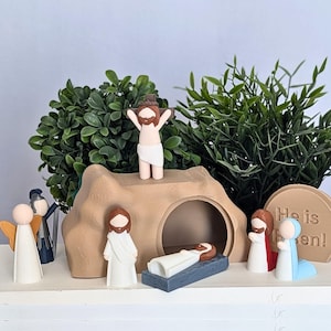 May include: A miniature nativity scene set with a brown cave, a white angel, a white figure of Jesus on the cross, a white figure of Jesus in the tomb, and a white figure of a woman with a blue robe. The scene is set on a white surface with green plants in the background. The text "He is risen!" is on a brown sign.