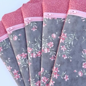 May include: A collection of fabric pouches with a floral pattern. The pouches have a gray background with pink flowers and green stems. The top of each pouch is a solid pink fabric with a pink zipper.