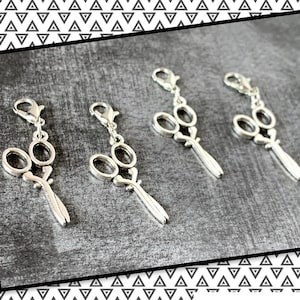 May include: Four silver scissor charms with a loop for attaching to a zipper pull or bag. The charms are detailed with a pair of scissors with a loop handle.